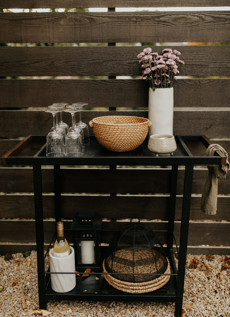 A bar cart sits in the rocks in a backyard, up against a wooden fence. The top of the bar cart has wine glasses, a candle, a basket and flowers, and the bottom has chargers and a bottle of wine chilling.