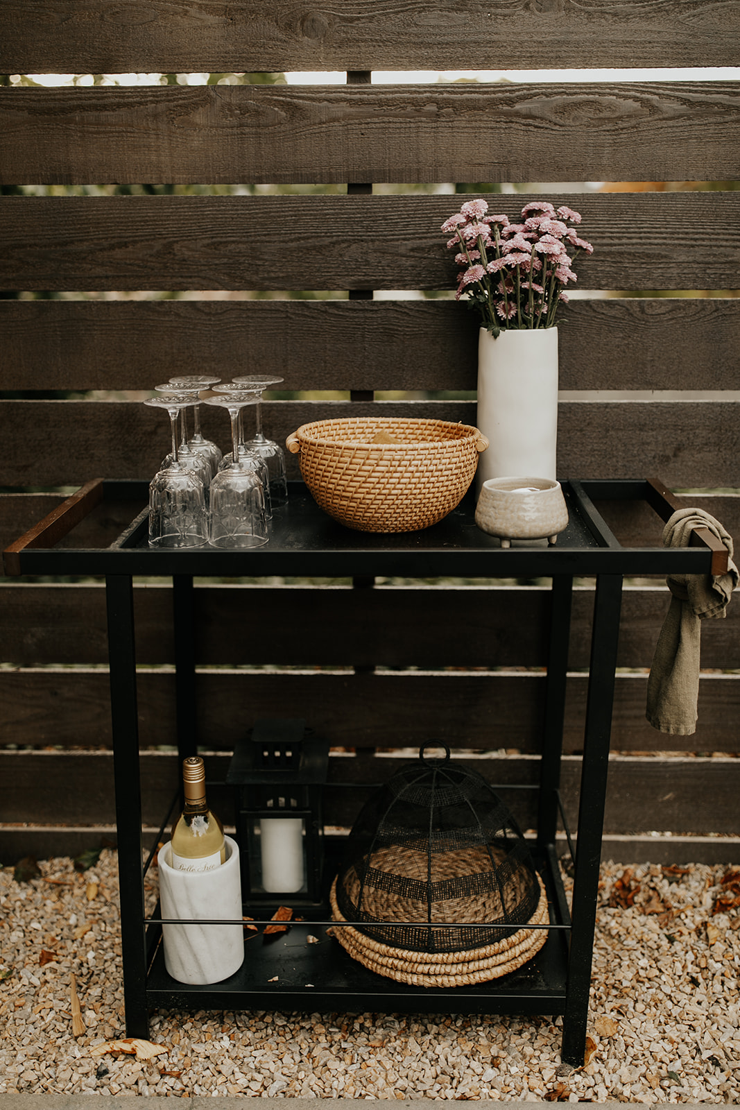 A bar cart sits in the rocks in a backyard, up against a wooden fence. The top of the bar cart has wine glasses, a candle, a basket and flowers, and the bottom has chargers and a bottle of wine chilling.