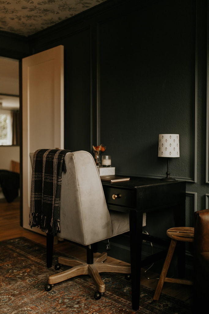 A black office desk sits in a room with blue-gray walls, a red rug, and wainscotting on the walls. A large comfortable office chair sits in front of the desk.