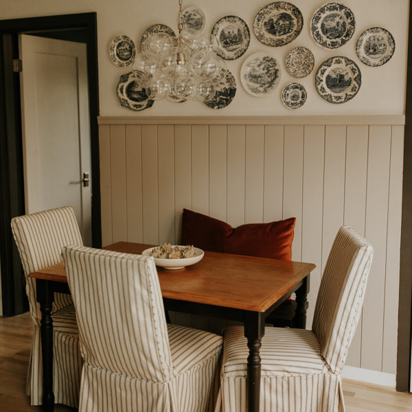 A dining room table sits on hardwood floors, with chairs on all sides, each of which has a striped slipcover on. The wall behind the table has partial shiplap and blue nordic plates hanging.