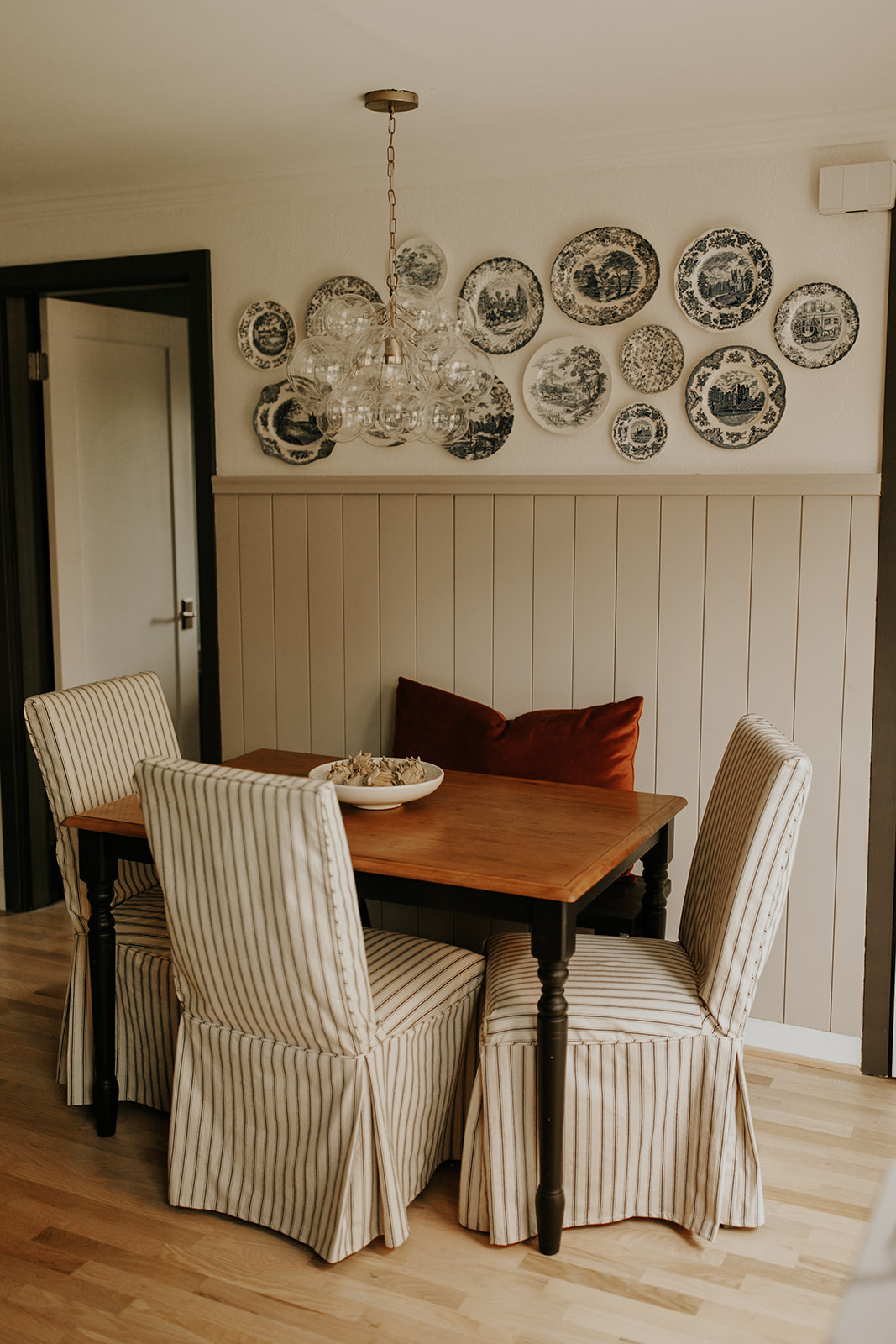 A dining room table sits on hardwood floors, with chairs on all sides, each of which has a striped slipcover on. The wall behind the table has partial shiplap and blue nordic plates hanging.