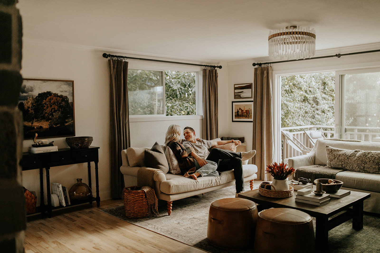 A man and woman sit on the couch in their living room, surrounded by a well decorated space complete with curtains, art, pillows, coffee table and hardwood floors