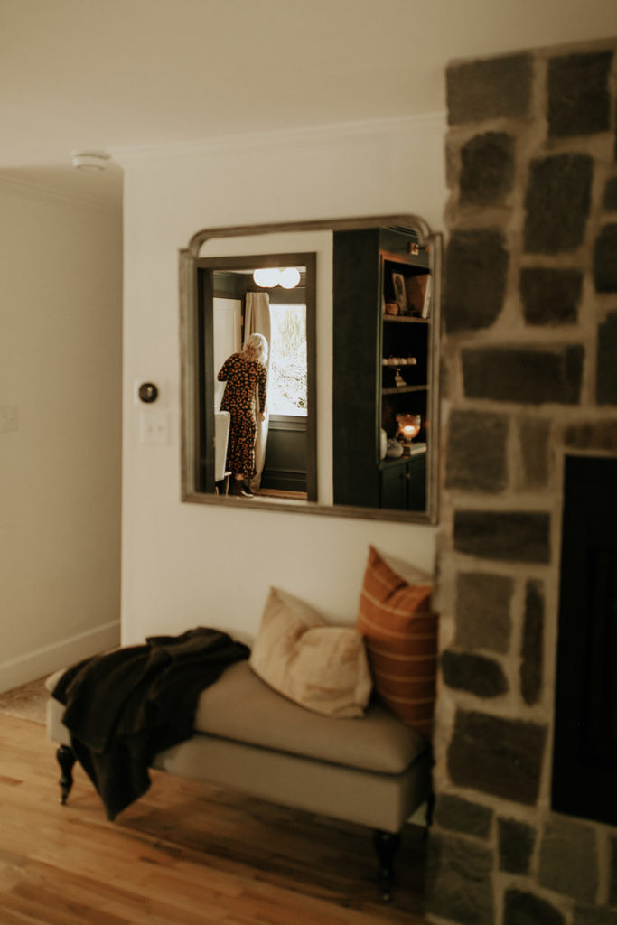 A mirror hangs on a wall next to a fireplace. In the reflection of a mirror, a woman wearing a dress stands in the next room fixing the curtains by the window