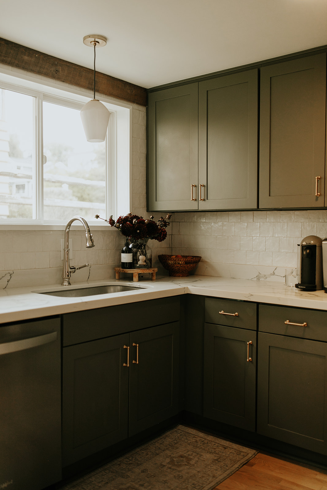 A photo of a kitchen with a wood beam on the wall, green kitchen cabinets with brass handles, a quartz countertop with stainless steel faucet and coffee maker on the counter.